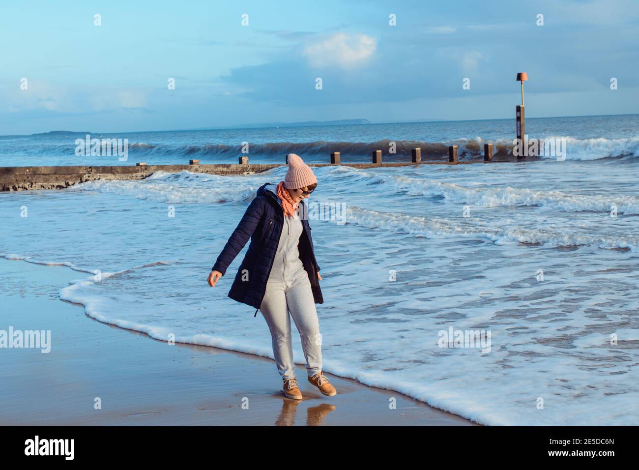 Femme en vêtements chauds et lunettes de soleil jouant avec les vagues au bord de la mer, profitant du moment pendant une promenade sur la côte. Détente et accomplissement personnel. Déplacement Banque D'Images