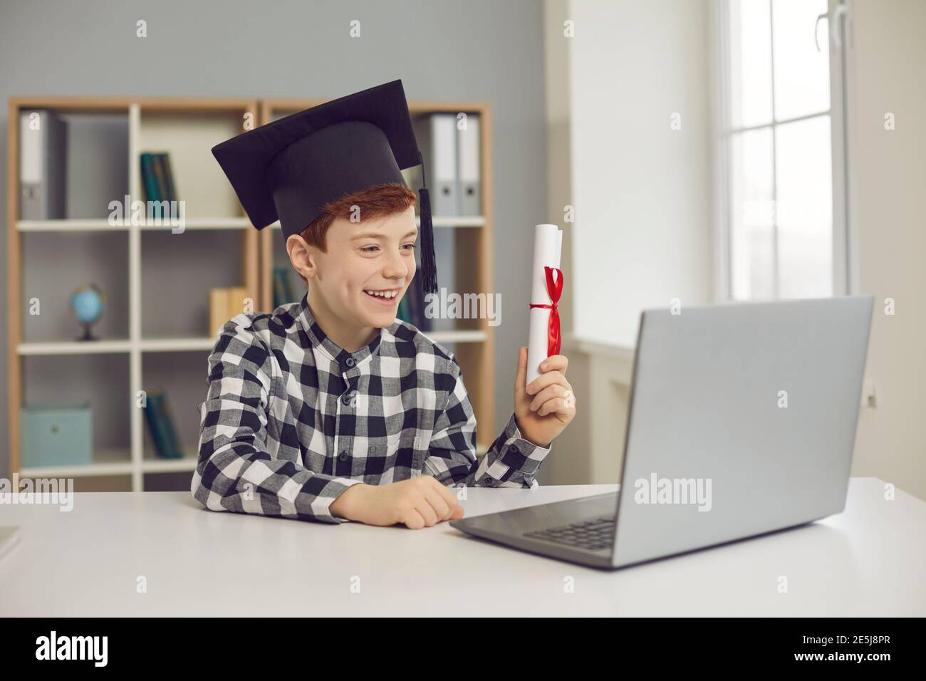 Garçon diplômé assis à un bureau dans un chapeau de troisième cycle et tenant un rouleau de diplôme devant un ordinateur portable. Banque D'Images