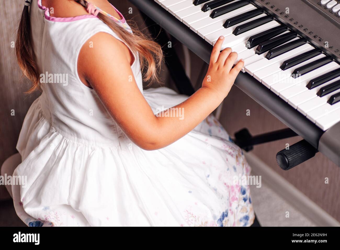 Vue arrière une fille dans une robe blanche d'été joue un synthétiseur électronique dans un intérieur de maison Banque D'Images