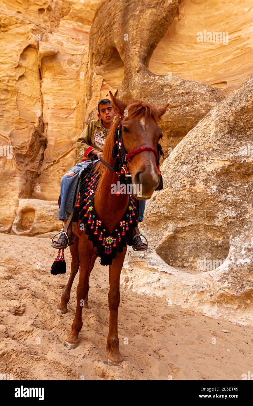 Petra, Jordanie 04-02-2010: Un garçon bédouin local portant des jeans et des sandales est sur un cheval avec un embout décoratif à la bouche. Ils se reposent à l'ombre d'une falaise Banque D'Images