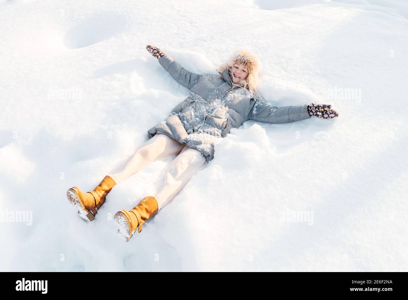 Belle souriante femme caucasienne adulte de taille moyenne portant une veste en duvet grise et des bottes jaunes couchés dans la neige faisant de l'ange de la neige. Banque D'Images