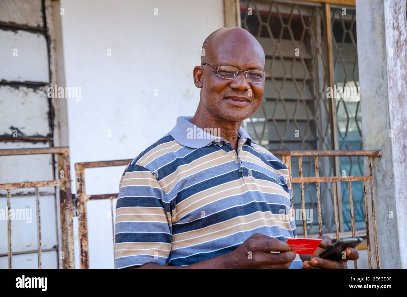 un vieil homme africain debout, utilisant son smartphone pour effectuer des paiements de certaines marchandises avec sa carte de débit/crédit Banque D'Images