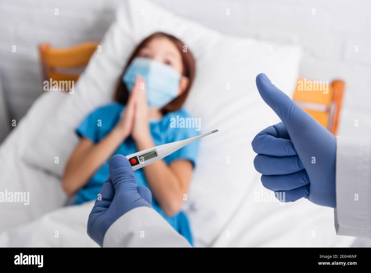 médecin tenant le thermomètre et montrant le pouce vers le haut près de l'enfant dans le masque médical allongé dans le lit avec les mains en prière, arrière-plan flou Banque D'Images