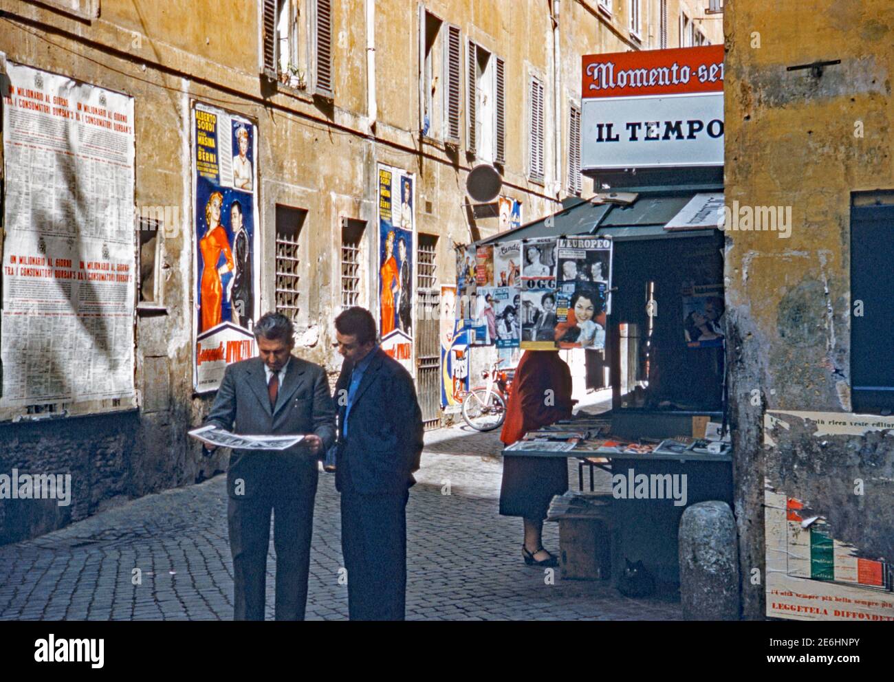 Un kiosque qui vend des magazines et des journaux dans les ruelles de Rome, en Italie, en 1955. Deux hommes parcourent les dernières nouvelles. Le signe au-dessus de la stalle annonce «il Tempo» (signifiant «heure» en anglais), un quotidien local. Les affiches sur le mur en face comprennent une pour le film «Buonanotte... Avvocato ! («Goodnight... Lawyer!»), un film humoristique italien réalisé par Giorgio Bianchi, avec Alberto Sordi, Maria Berni et Giulietta Masina et sorti cette année-là. Banque D'Images
