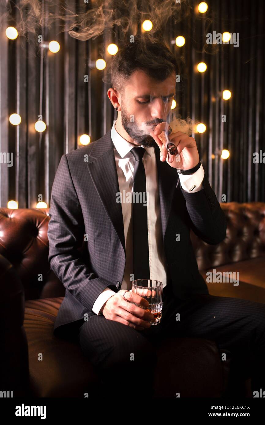 Portrait d'un homme brutal de barbe avec tatouage fumant cigare dans chaud élégant costume boire l'alcool de whisky dans l'appartement loft . Couple Love Story concept Banque D'Images