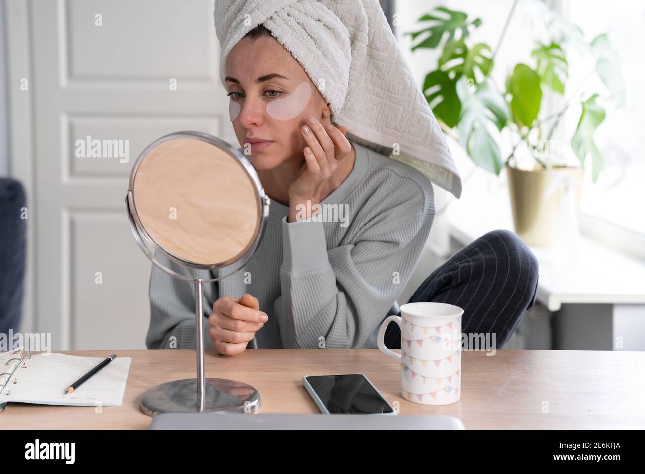 Femme avec une serviette sur la tête appliquer des plaques de récupération sous les yeux d'hydrogel à la maison, regardant dans le miroir. Banque D'Images