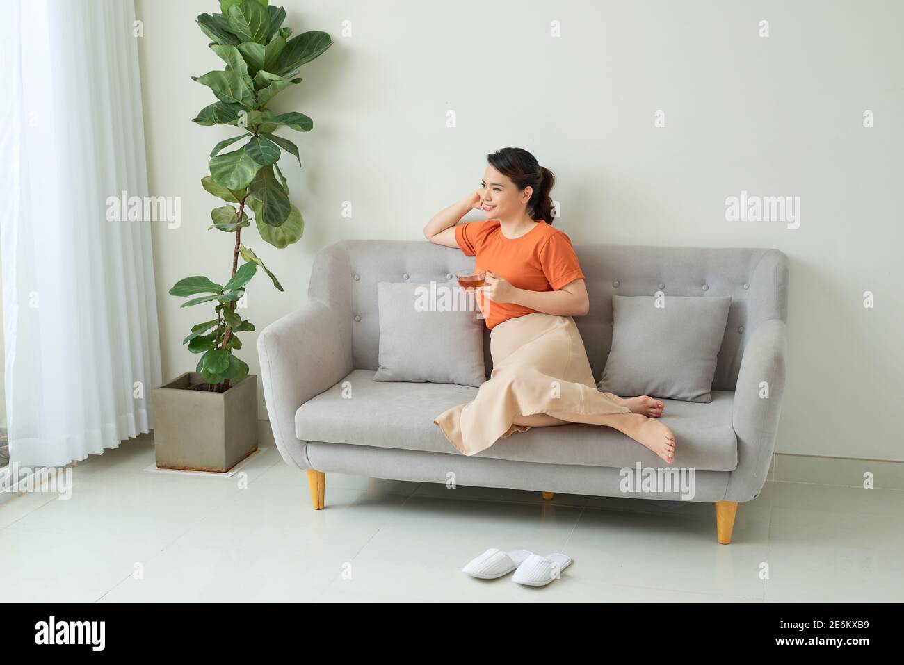 Jeune femme souriante assise sur un canapé et regardant vers le haut tout en buvant du thé chaud. Banque D'Images