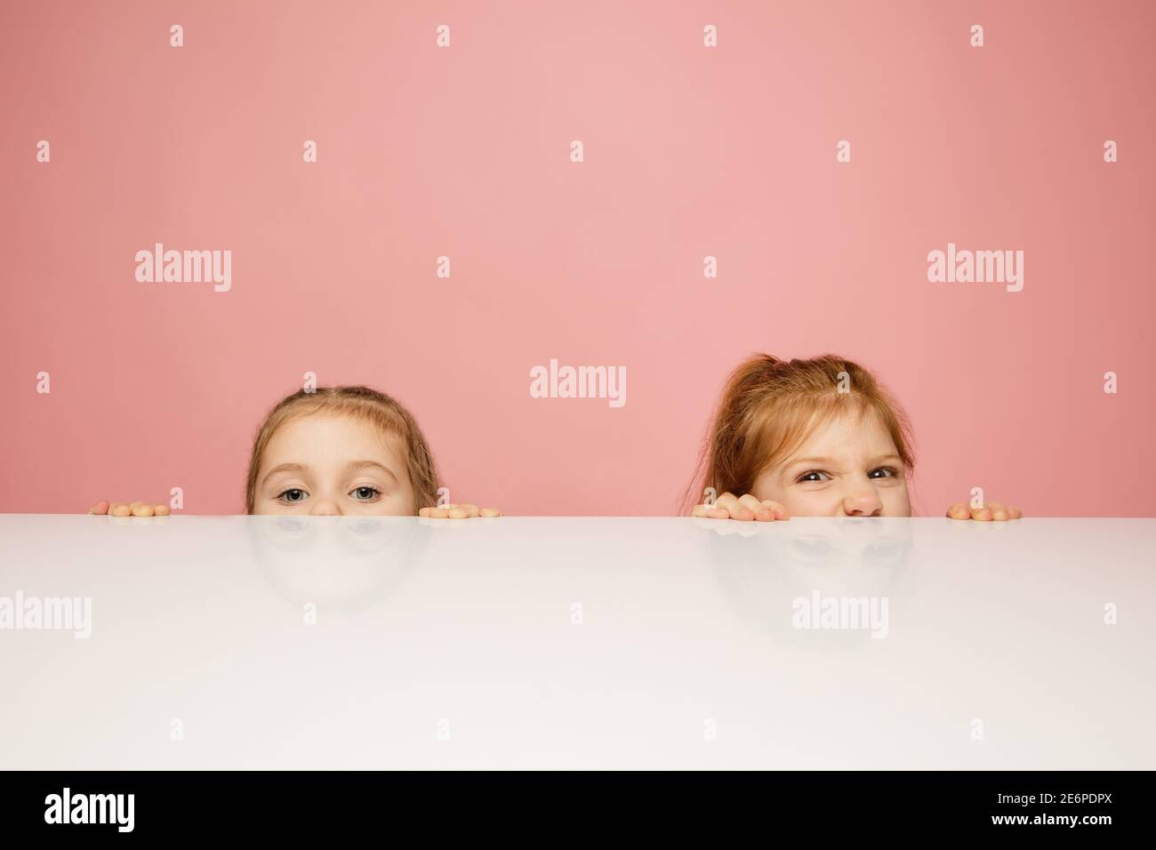 Masquage. Enfants heureux, filles isolées sur fond de studio rose corail. Vous pouvez être heureux, joyeux. CopySpace pour publicité. Enfance, éducation, émotions, concept d'expression faciale. En partant de l'arrière de la table. Banque D'Images