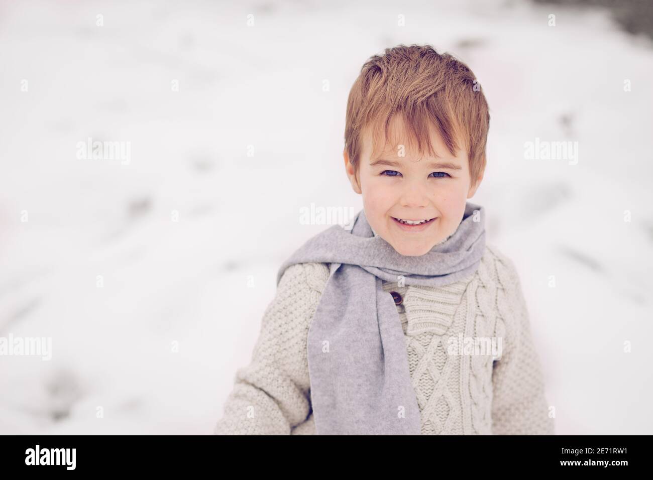 Petite blonde blanc garçon enfant tout-petit dans la neige d'hiver portant écharpe et chandail riant et regardant gentille. Banque D'Images