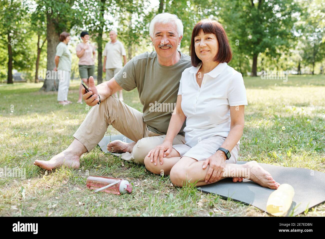 Portrait d'un couple senior regardant l'appareil photo tout en étant assis herbe verte et exercice dans le parc Banque D'Images