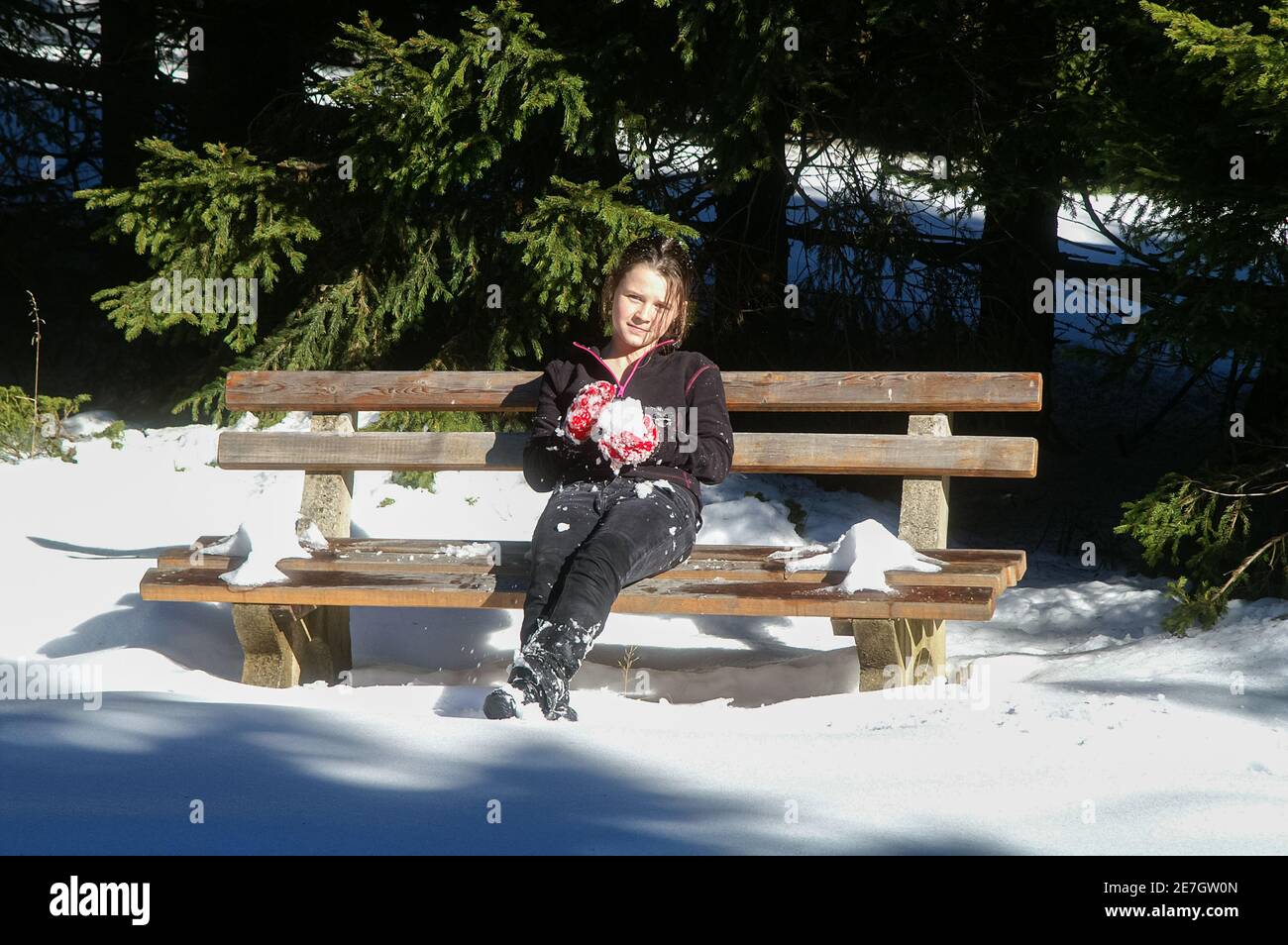 Une jeune fille dans une forêt enneigée d'hiver dans un survêtement noir, assise sur un banc faisant un boule de neige Banque D'Images