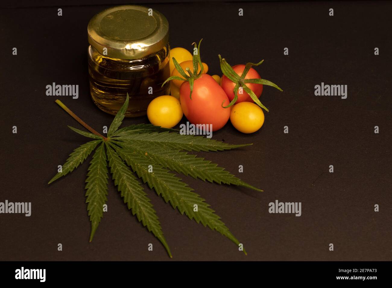 Une feuille de cannabis, un pot rempli d'huile de cbd et quelques tomates cerises sur fond noir. L'image illustre la relation entre l'alimentation et le cannabis. Banque D'Images