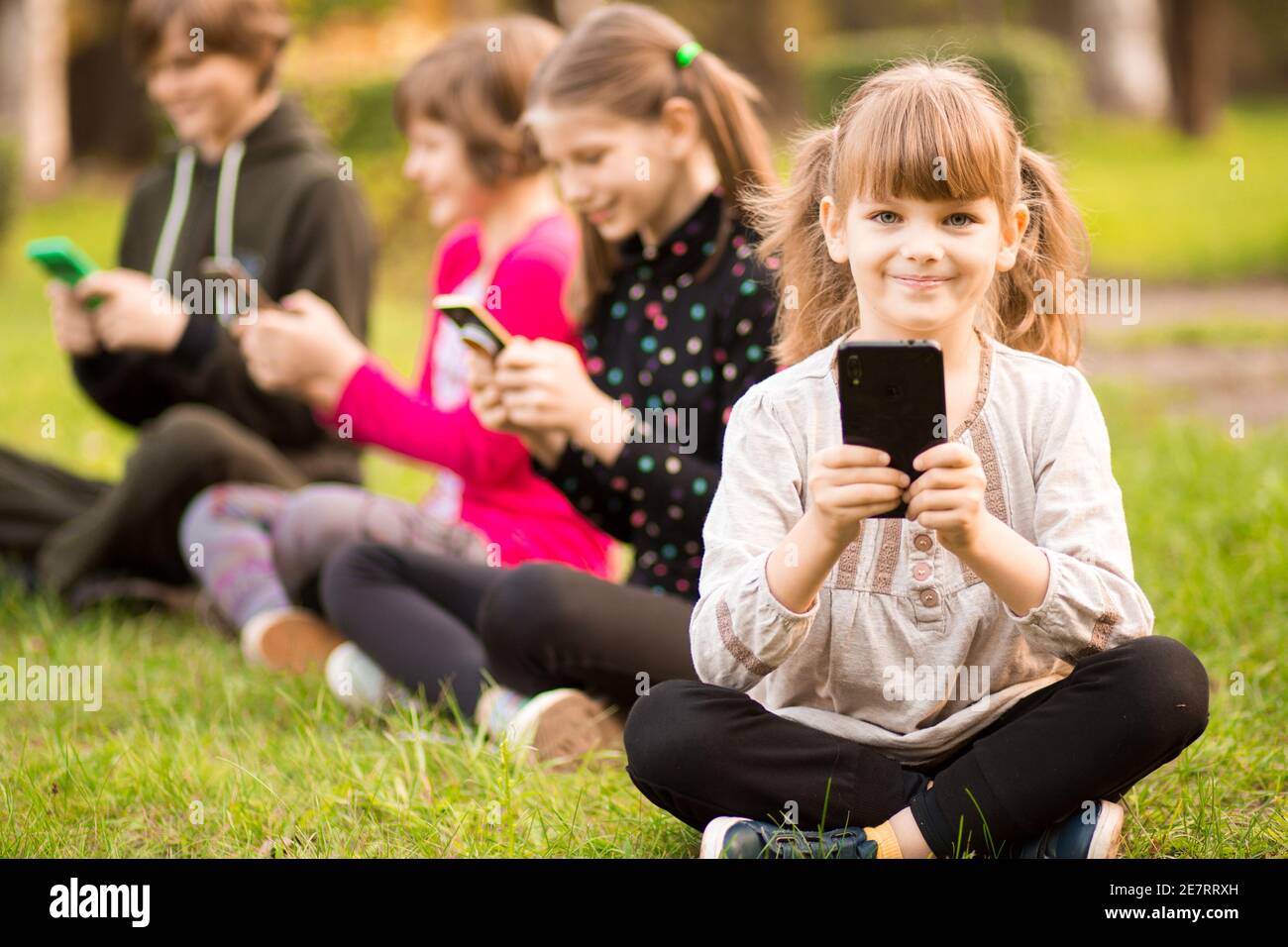 Photo pleine longueur d'un portrait extérieur d'enfant positif inspiré, fille assise sur l'herbe avec les jambes croisées et en utilisant le téléphone portable en compagnie d'amis Banque D'Images