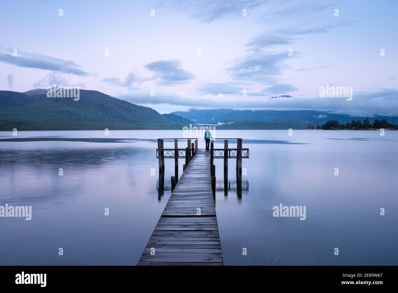 Femme solitaire debout au bout de la jetée du lac te Anau au lever du soleil, regardant les montagnes Kepler. Format paysage. Banque D'Images