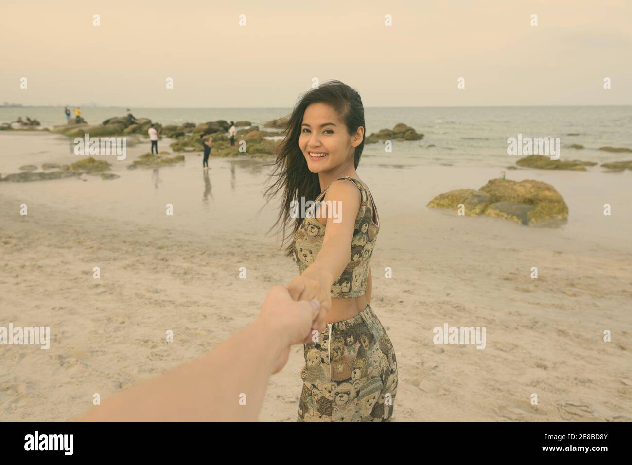 Young happy Asian woman smiling while holding hands and leading man à la plage publique de Hua Hin en Thaïlande Banque D'Images