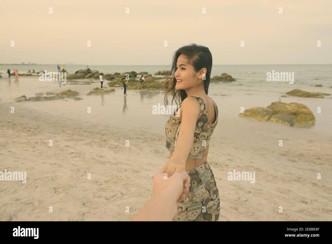 Young happy Asian woman smiling while holding hands and leading man à la plage publique de Hua Hin en Thaïlande Banque D'Images