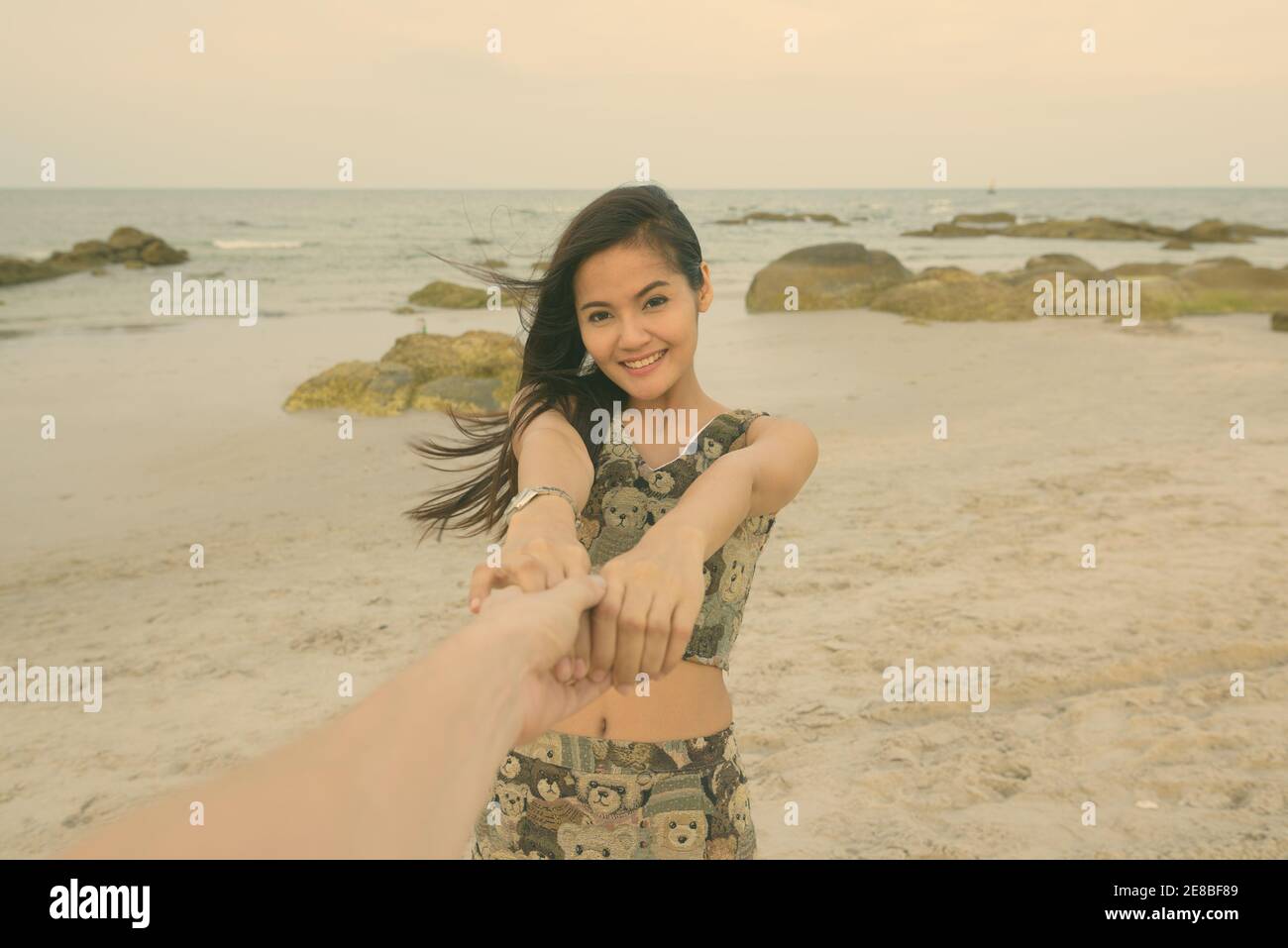 Young happy Asian woman smiling while holding avec les deux mains et l'homme à la plage publique de Hua Hin en Thaïlande Banque D'Images