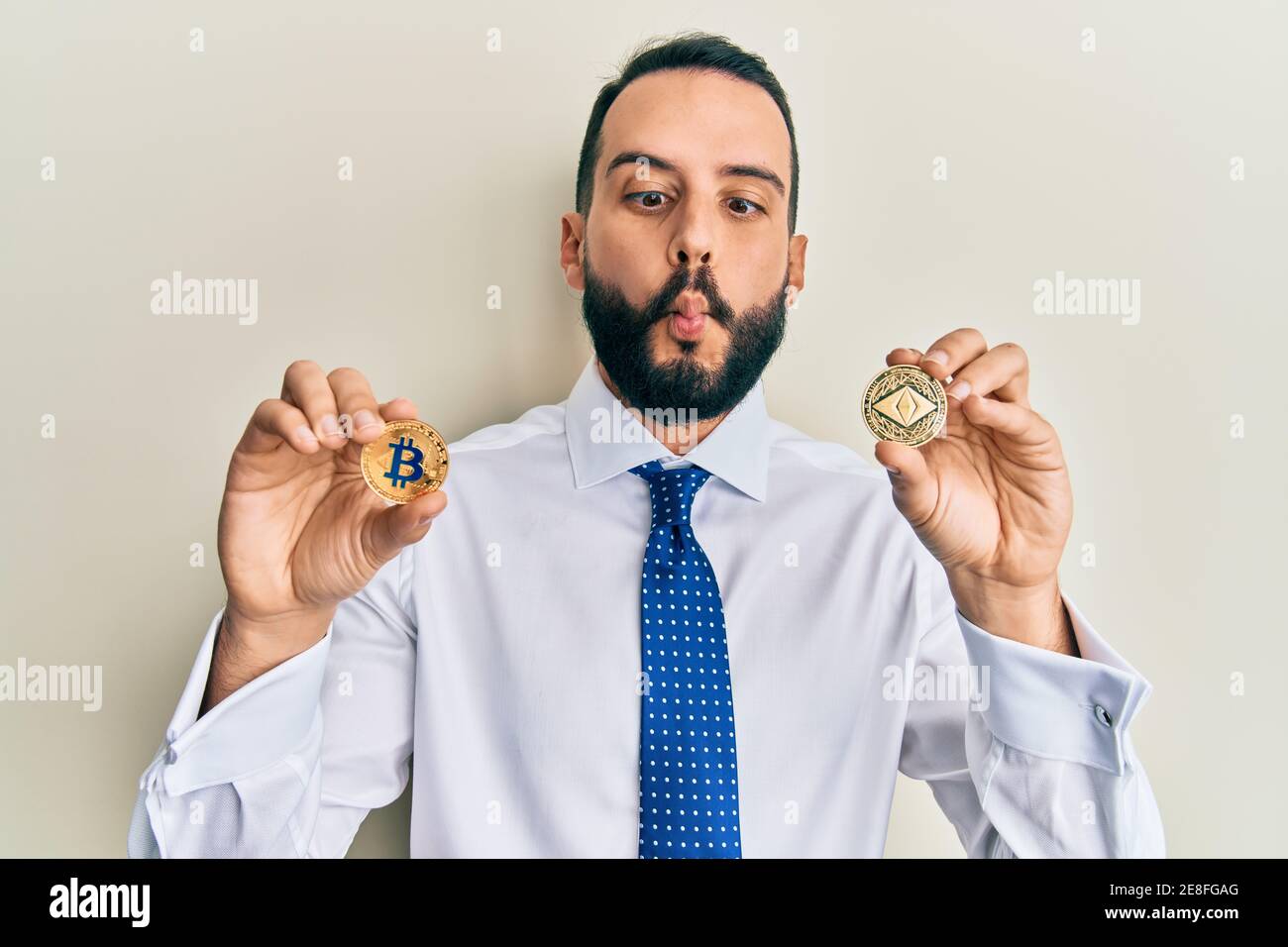 Jeune homme avec la barbe tenant la monnaie virtuelle éthereum pièce et bitcoin faisant le poisson visage avec la bouche et les yeux de bourdonnement, fou et comique. Banque D'Images