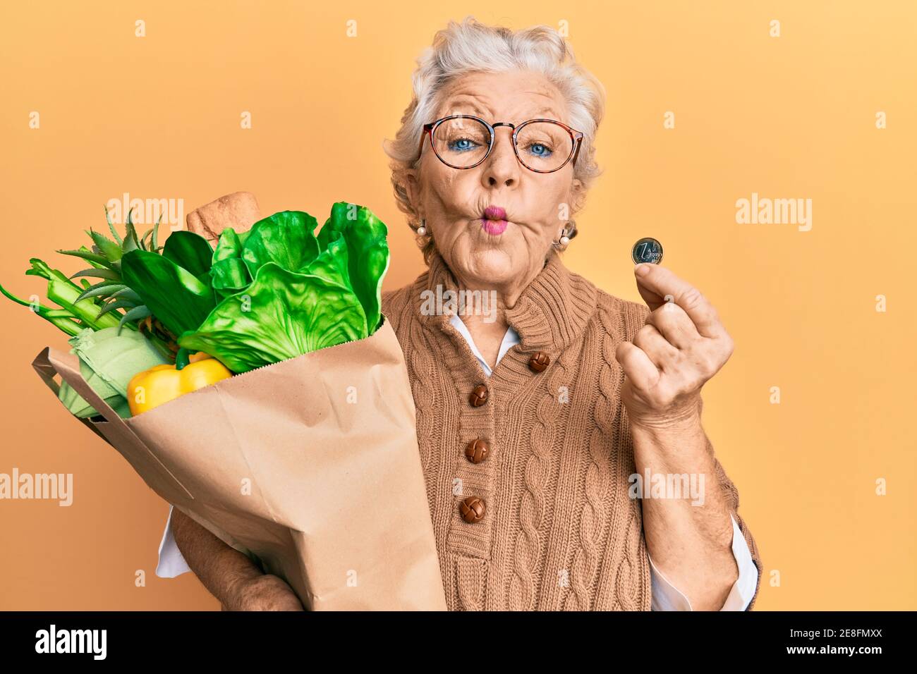 Femme sénior aux cheveux gris tenant des épiceries et 1 euro de pièce de fabrication de poisson visage avec la bouche et les yeux accroupidés, fou et comique. Banque D'Images