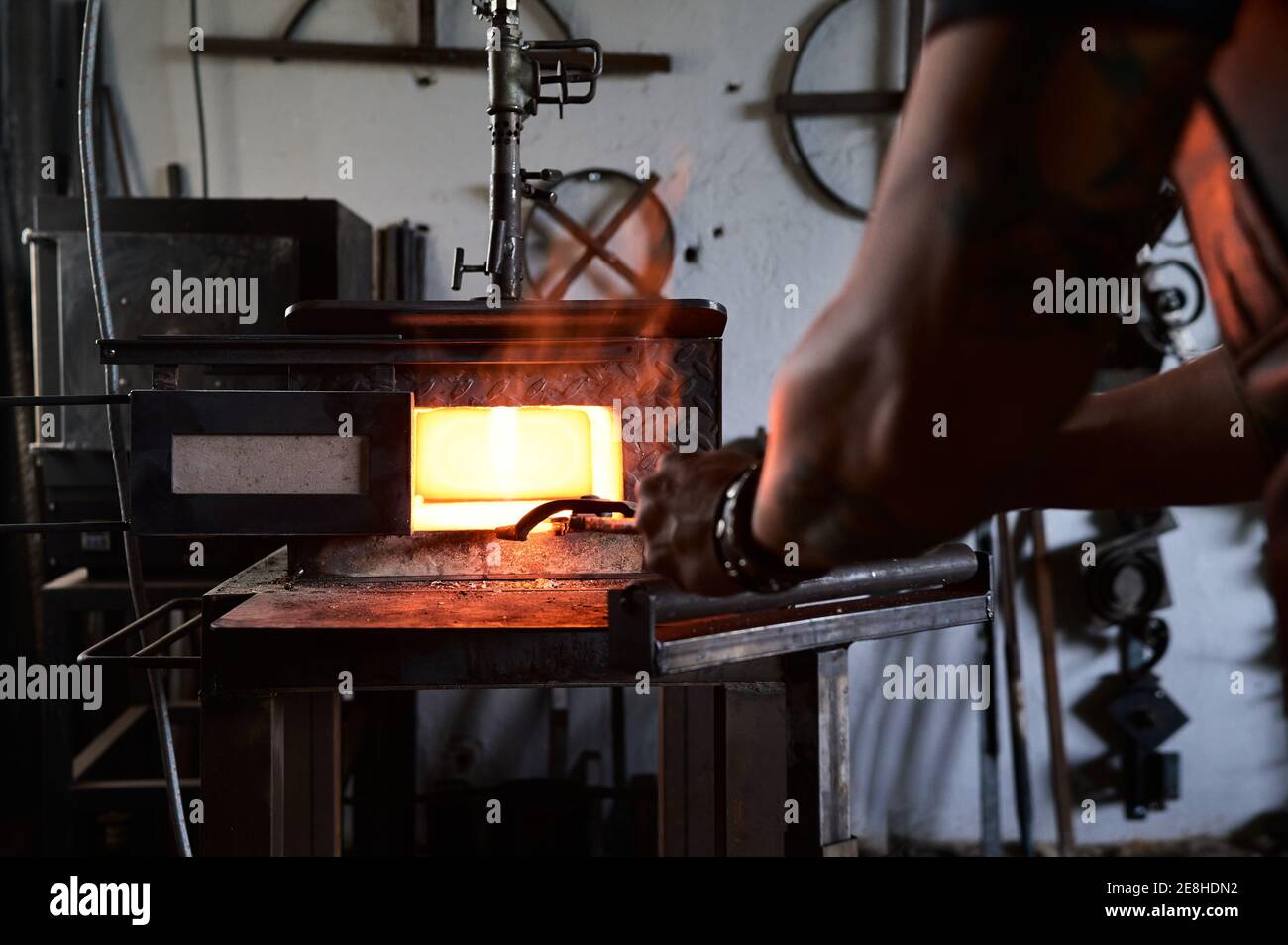 Vue arrière du jeune homme tatoué anonyme en tablier chauffer du métal dans un four tout en travaillant dans des taches de terre Banque D'Images Vue arrière du jeune homme tatoué anonyme en tablier chauffer du métal dans un four tout en travaillant dans des taches de terre Banque D'Images