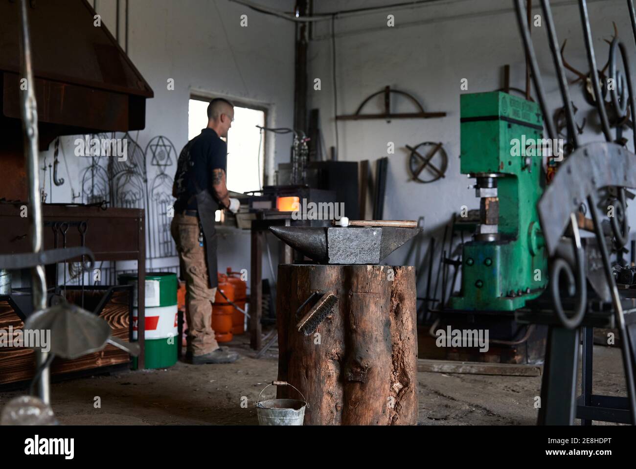 Vue arrière de la foreur de jeune homme sérieusement tatouée dans le tablier chauffer du métal dans un four tout en travaillant dans des taches de terre Banque D'Images Vue arrière de la foreur de jeune homme sérieusement tatouée dans le tablier chauffer du métal dans un four tout en travaillant dans des taches de terre Banque D'Images