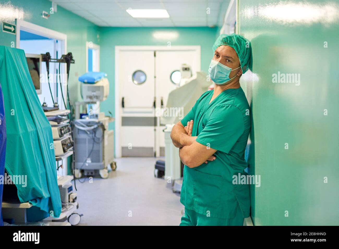 Vue latérale de jeune homme fatigué médecin dans les exfoliants et masque médical reposant sur le mur avec les bras pliés au repos dans le couloir de l'hôpital après la chirurgie Banque D'Images