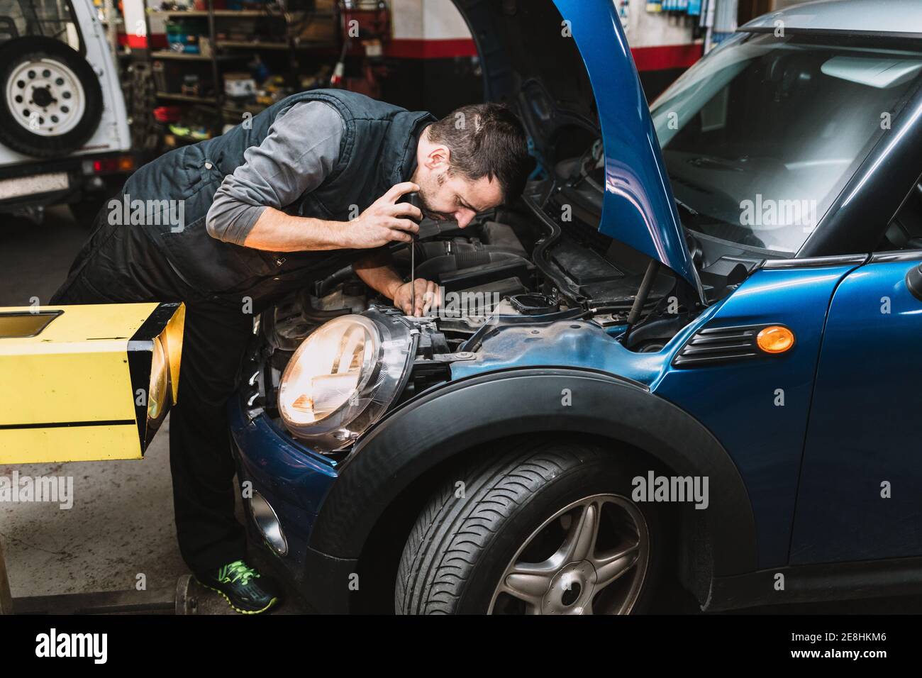 Jeune homme d'entretien barbu concentré répare la voiture contemporaine contre Bright lumière en atelier Banque D'Images