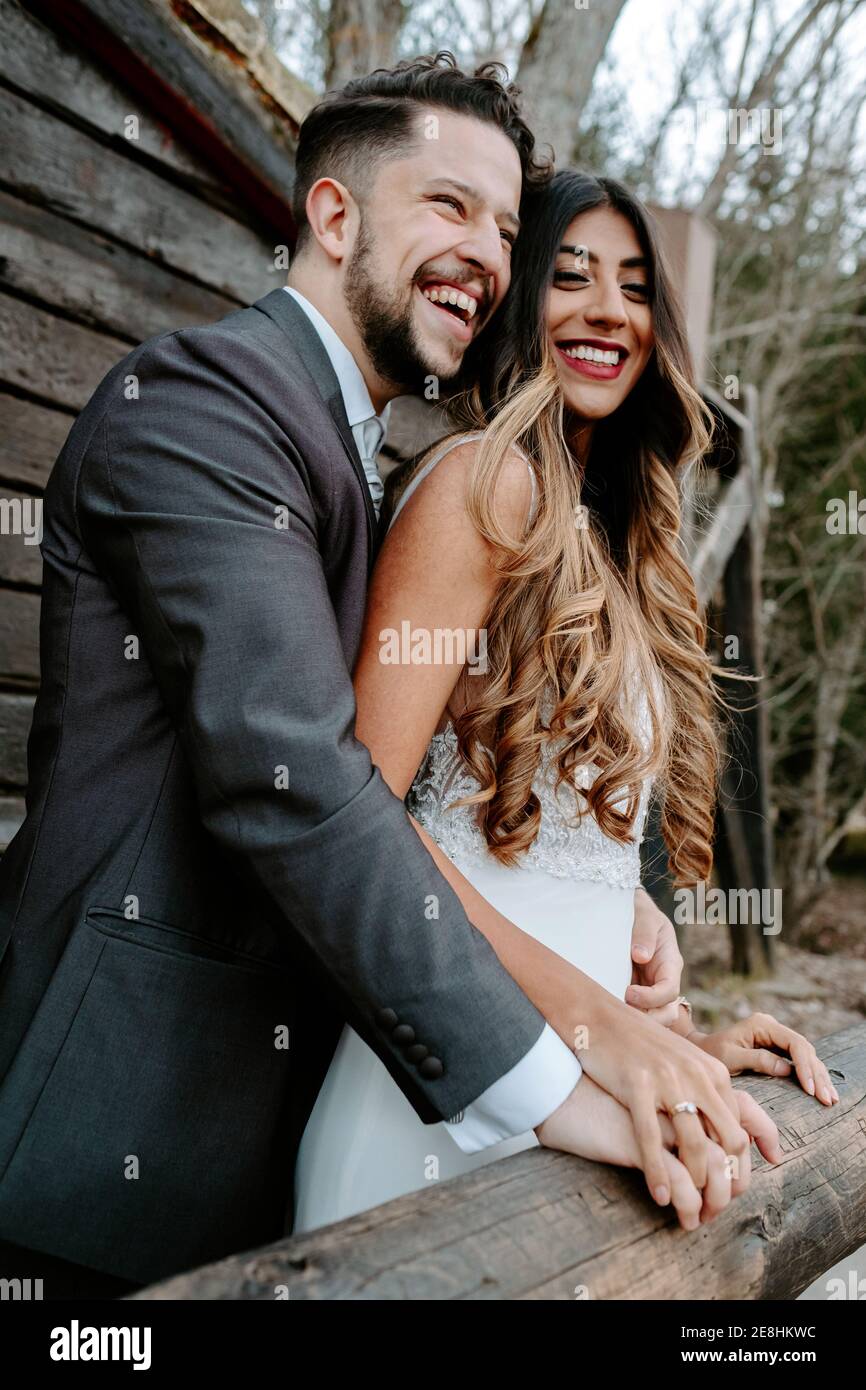 Vue latérale d'un jeune gai à barbe en costume riant et une élégante mariée ethnique en robe blanche tout en étant debout ensemble sur pont en bois n Banque D'Images