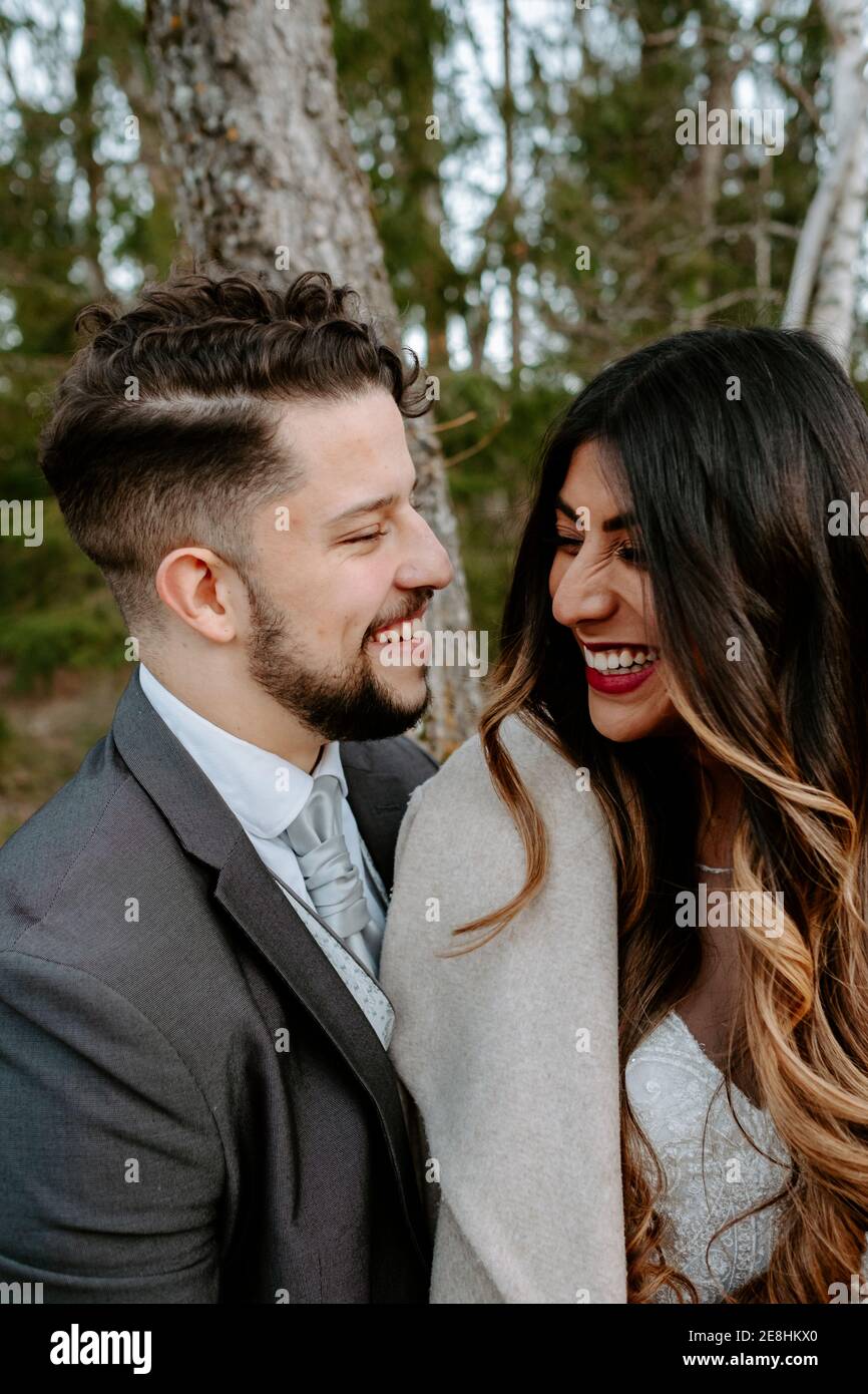 Vue latérale d'un jeune gai à barbe en costume riant et une élégante mariée ethnique en robe blanche tout en étant debout ensemble sur pont en bois n Banque D'Images