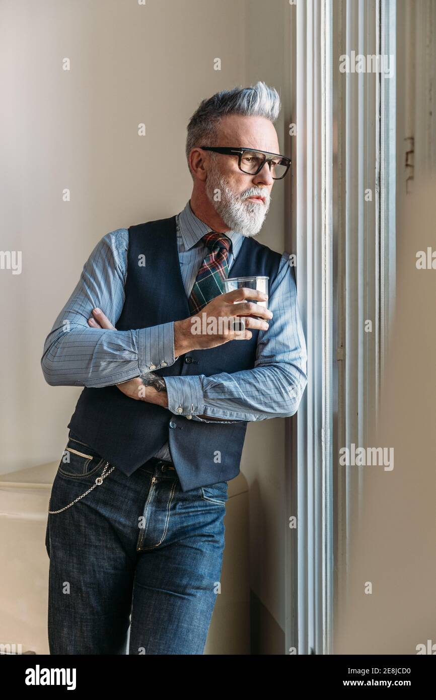 Homme d'âge moyen plein de confiance avec cheveux et barbe gris dans une  tenue tendance et des lunettes penchées sur le mur et regardant par la  fenêtre pendant que vous buvez du
