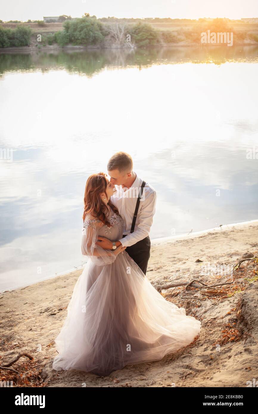 Un beau couple de mariage sur la rive au coucher du soleil. Une femme dans  une robe grise avec des fleurs, un homme dans un costume et des bretelles  s'embrassent, l'amour. Amour