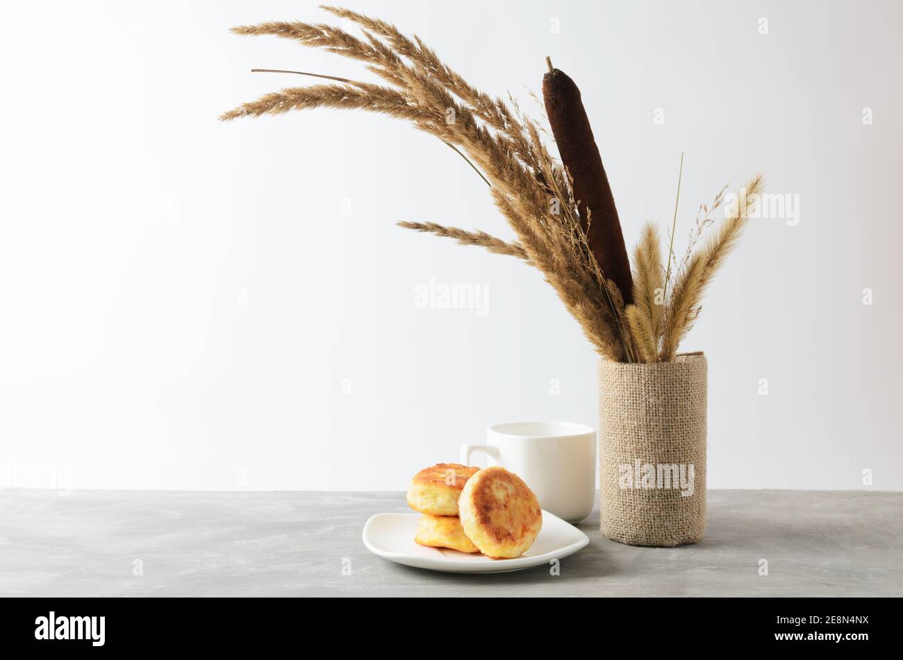 Quelques crêpes sur une assiette blanche et une tasse vide sur une table grise texturée contre un mur blanc avec un bouquet d'automne d'herbes séchées et de roseaux. Sélectionnez Banque D'Images