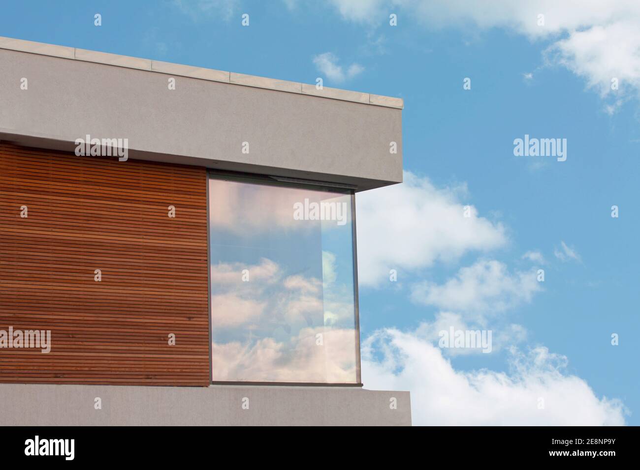 Détail d'une maison moderne en cube avec panneaux en bois et ciel bleu avec des nuages blancs reflétant dans les grandes fenêtres. Banque D'Images