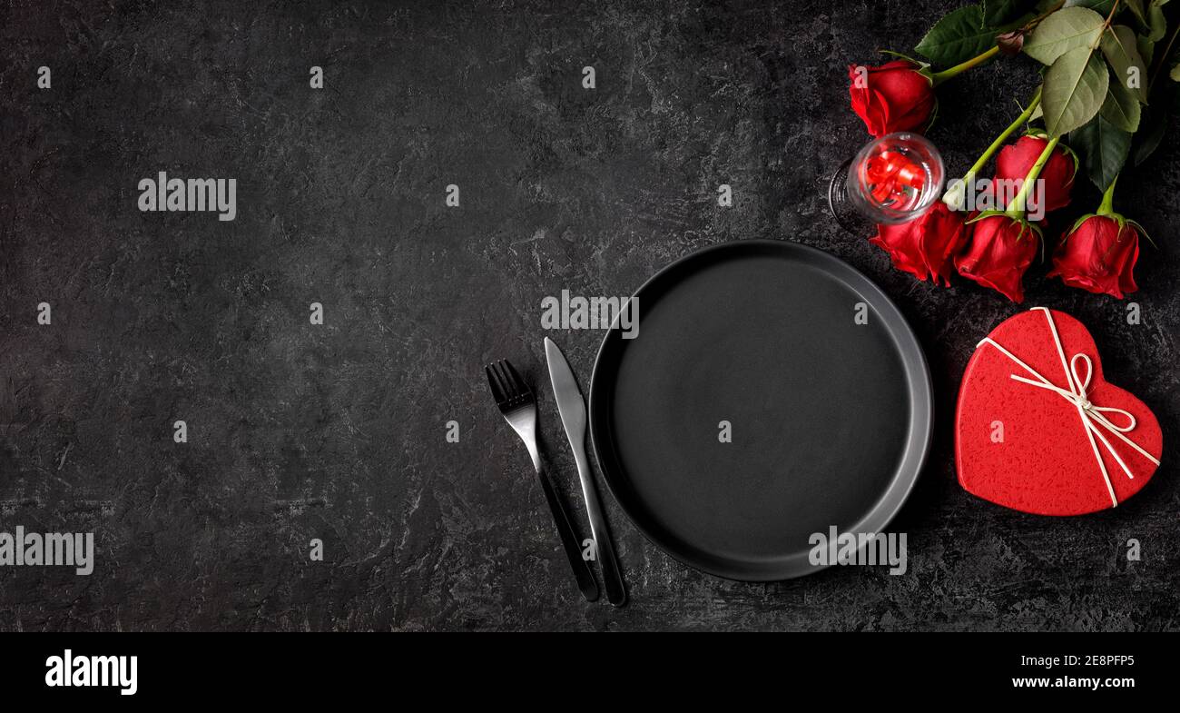 Dîner de fête de la Saint-Valentin ou du jour de naissance avec une assiette mate noire vide, une boîte cadeau en forme de foyer et des roses rouges Banque D'Images