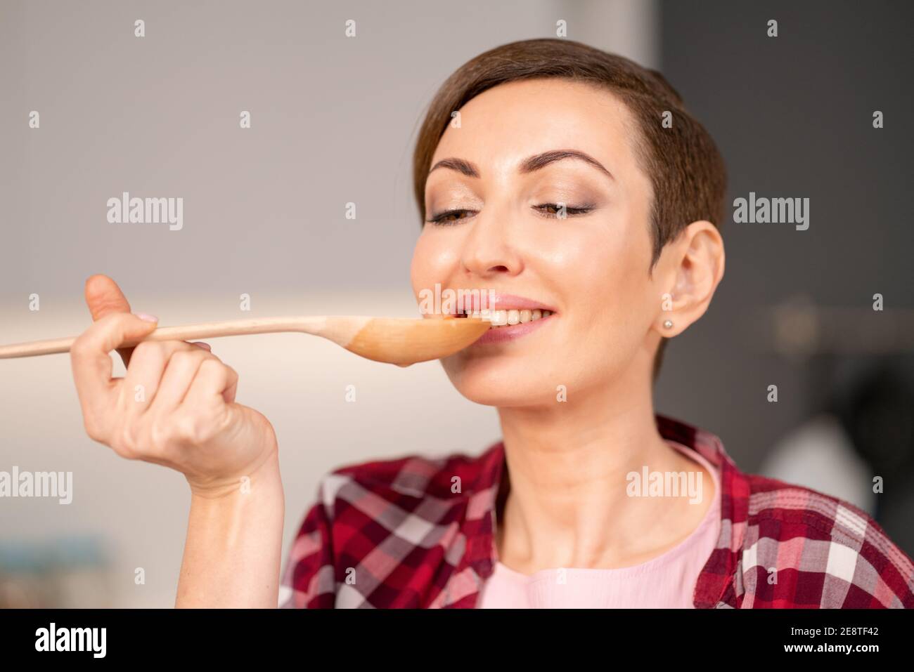 Gros plan d'une femme essayant un plat cuit à l'aide d'une longue cuillère en bois. Femme avec un cheveux court de cuisine un dîner pour la famille debout dans la cuisine moderne Banque D'Images