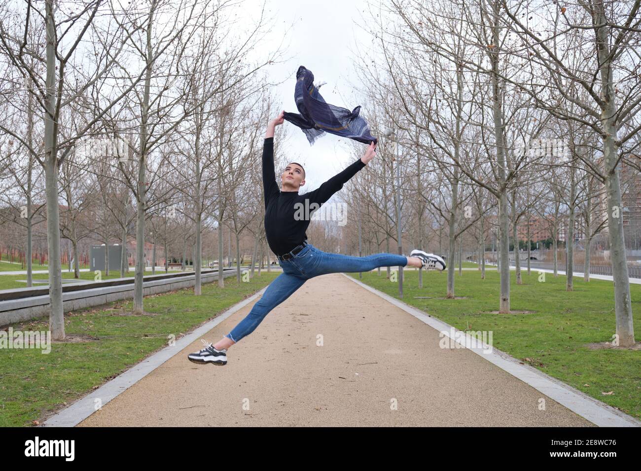 Beau jeune homme portant un maquillage et des écouteurs, faisant un saut de ballet dans un sentier de parc. Type androgyne non binaire. Banque D'Images