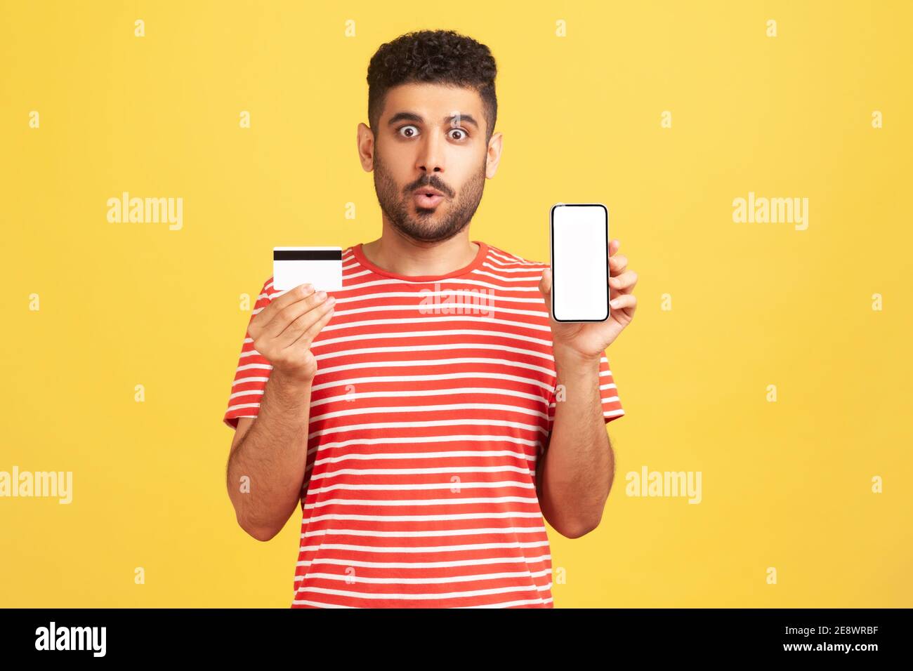 Un homme excité choqué avec une barbe en t-shirt rayé tenant une carte de crédit et un smartphone, regardant l'appareil photo avec de grands yeux, surpris par la facilité de banque. Po Banque D'Images