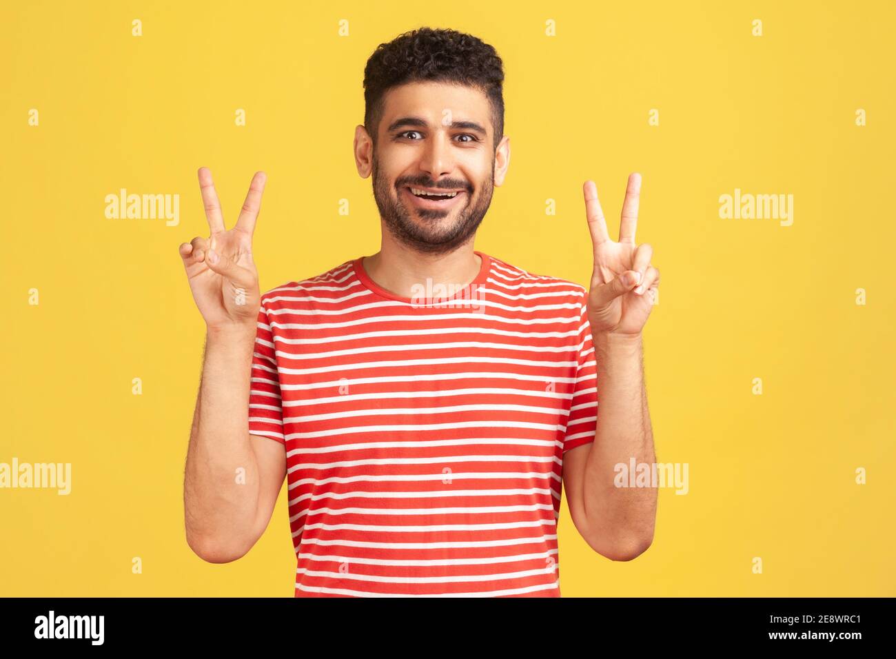Joyeux homme réussi avec une barbe en t-shirt rayé montrant le geste de victoire du signe v avec les doigts, regardant l'appareil photo avec un sourire crasseux. Studio intérieur sho Banque D'Images
