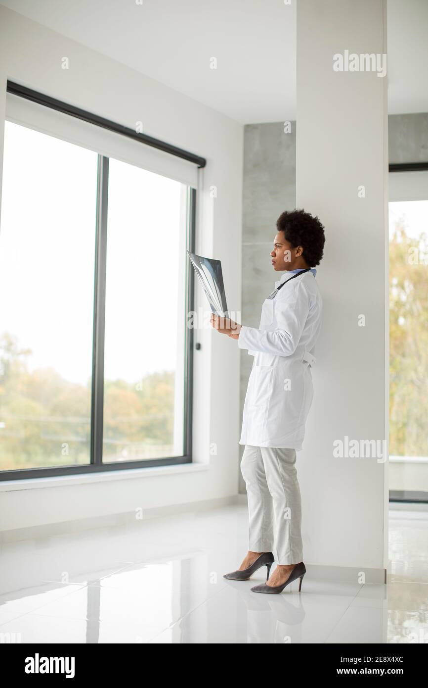 Femme afro-américaine médecin portant un manteau blanc avec stéthoscope debout par la fenêtre dans le bureau et regardant l'image de rayons x. Banque D'Images