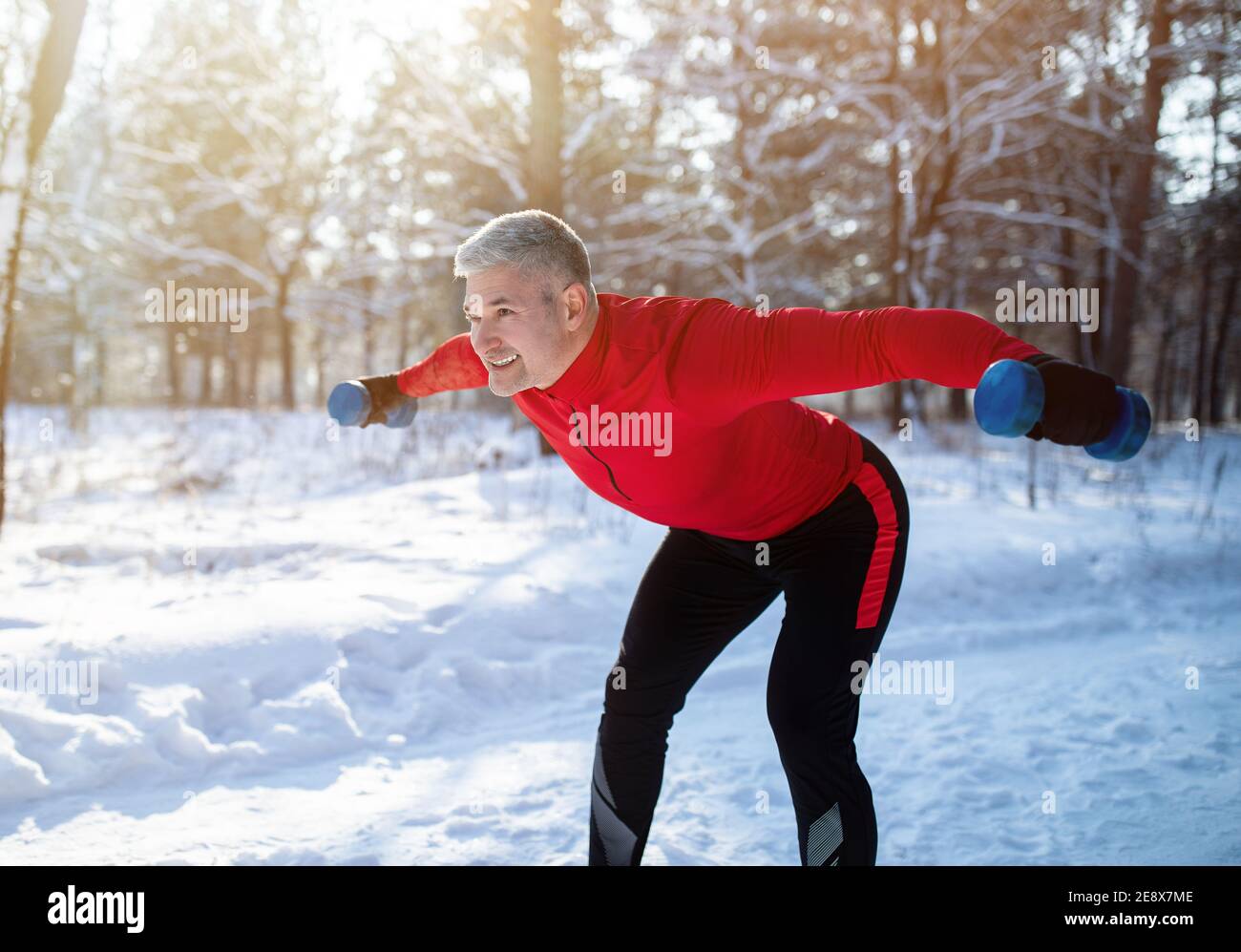 Entraînement de musculation en plein air en hiver. Sportif senior positif avec haltères effectuant des exercices de soulèvement dans un parc enneigé Banque D'Images