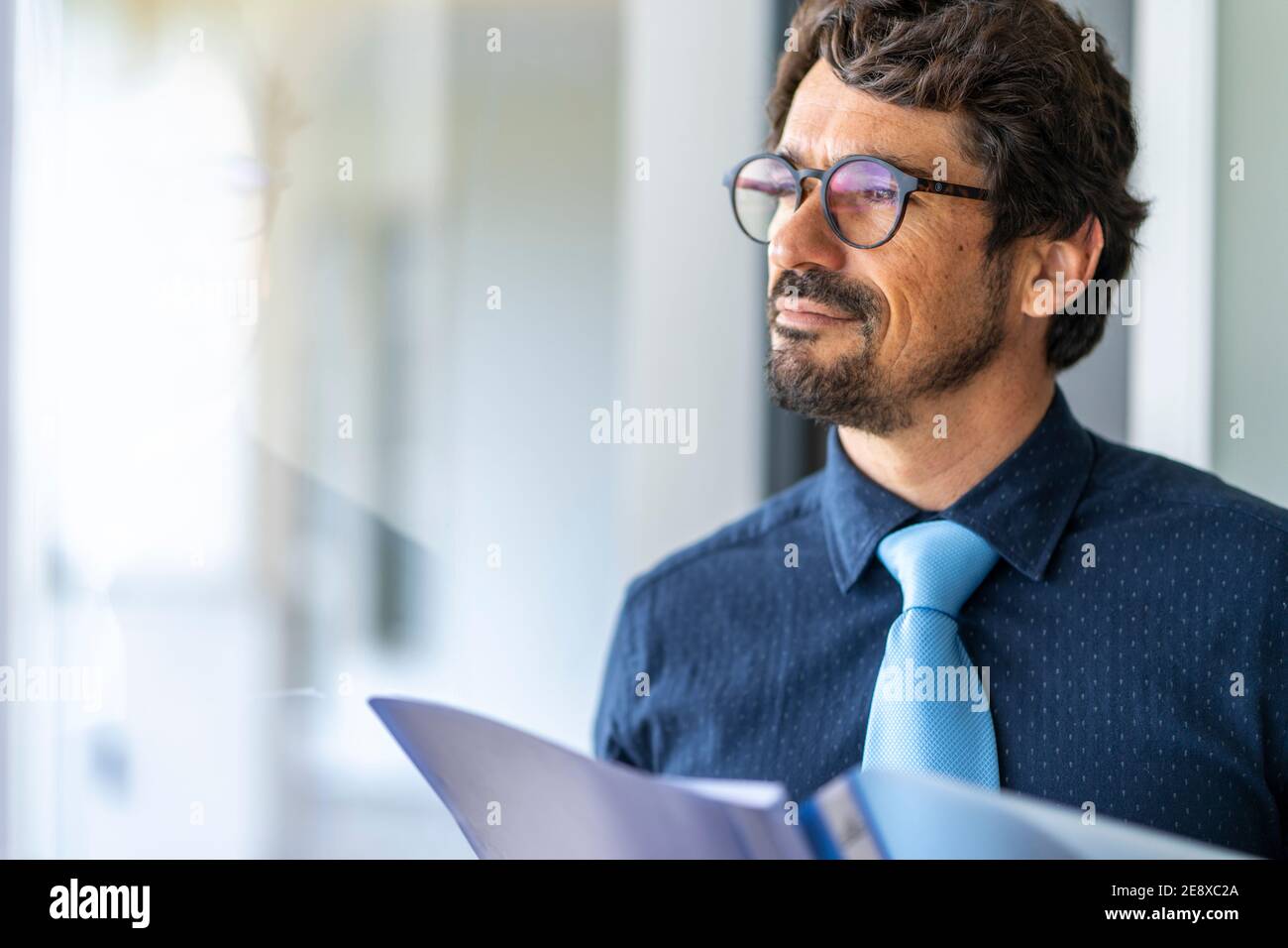 Homme d'affaires portant des lunettes tenant une chemise de documents en papier. Portrait d'homme réussi regardant par la feuille de contrat de fenêtre au bureau Banque D'Images