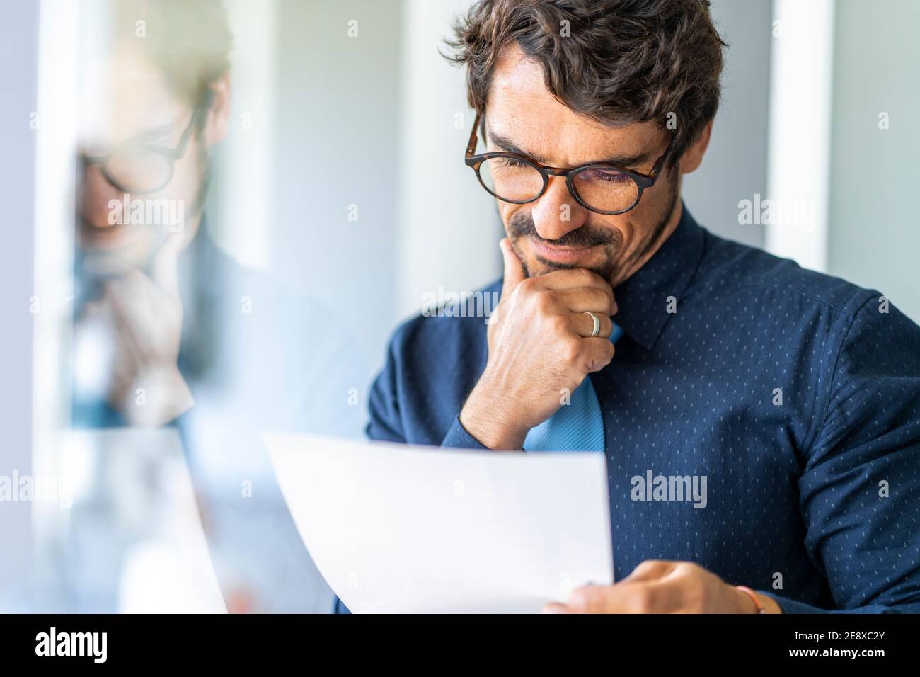 Homme d'affaires portant des lunettes tenant un document en papier. Contrat réussi de réflexion et de lecture de portrait masculin au bureau près de la fenêtre Banque D'Images