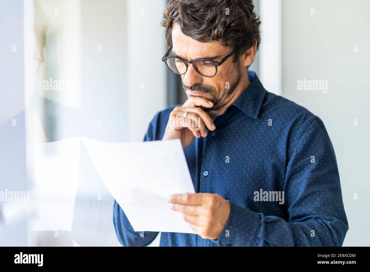 Homme d'affaires portant des lunettes tenant un document en papier. Contrat réussi de réflexion et de lecture de portrait masculin au bureau près de la fenêtre Banque D'Images