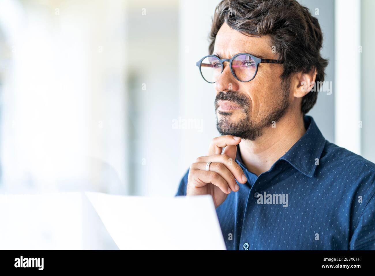 Homme d'affaires portant des lunettes tenant un document en papier regardant la fenêtre. Contrat réussi de réflexion et de lecture de portrait masculin au bureau Banque D'Images