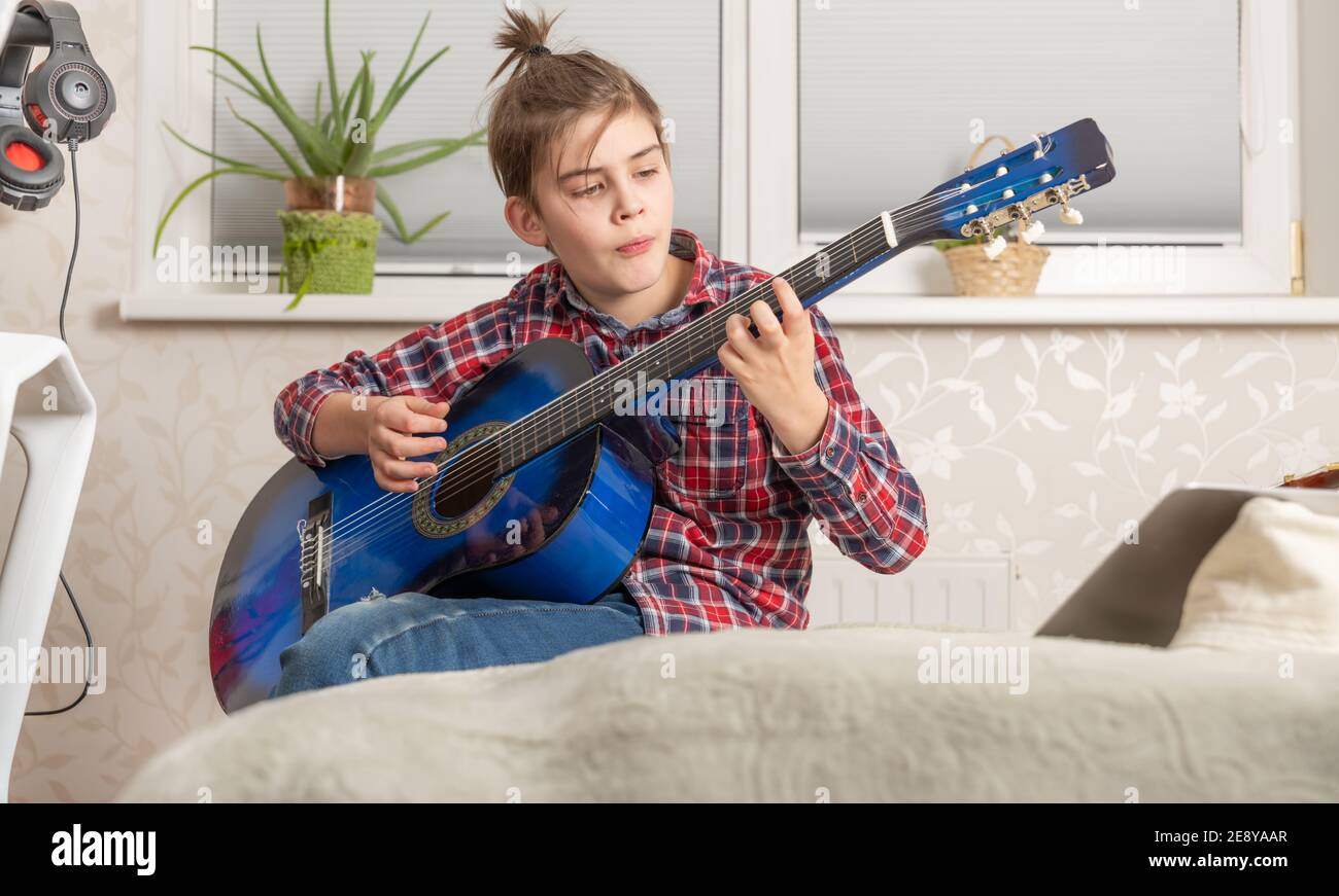 joyeux garçon adolescent en jeans et chemise rouge avec queue de cheval jouant de la guitare acoustique à la chambre à coucher, leçons de guitare et concept d'éducation musicale Banque D'Images