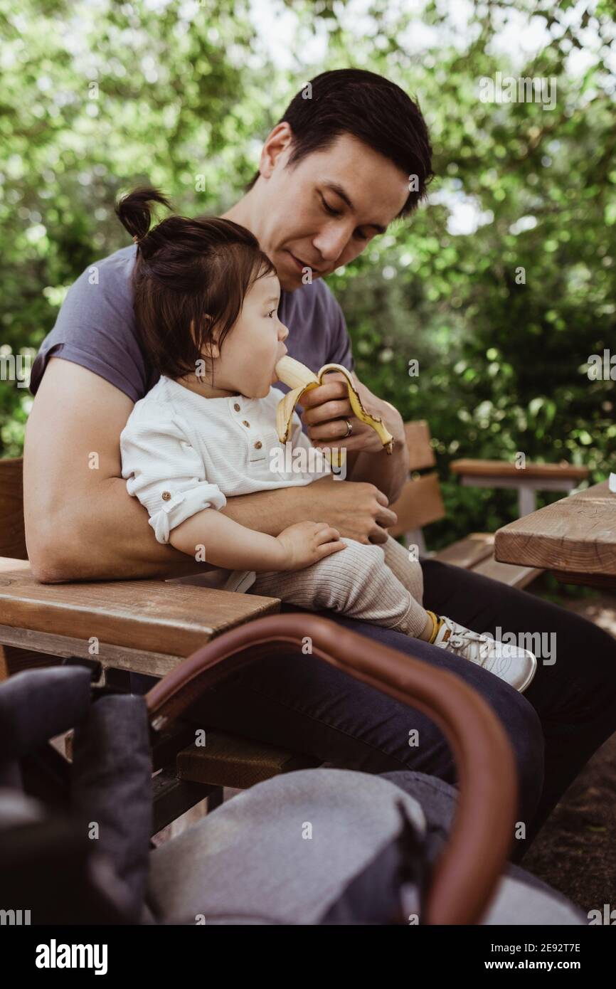 Père nourrissant de la banane à un enfant tout-petit tout en étant assis dans le parc Banque D'Images
