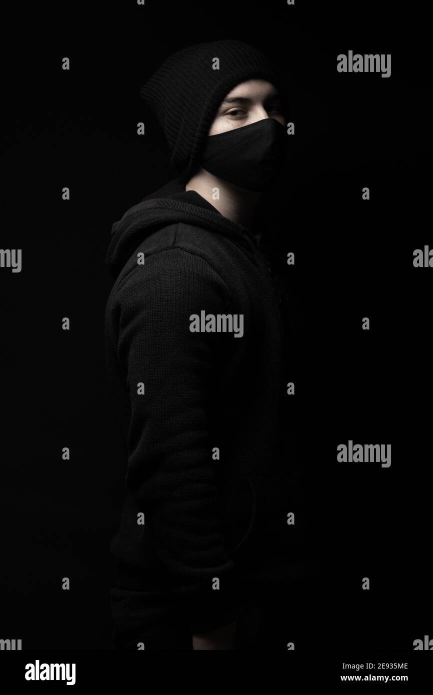 jeune homme avec bonnet en tricot d'hiver noir et masque facial debout devant un arrière-plan noir Banque D'Images