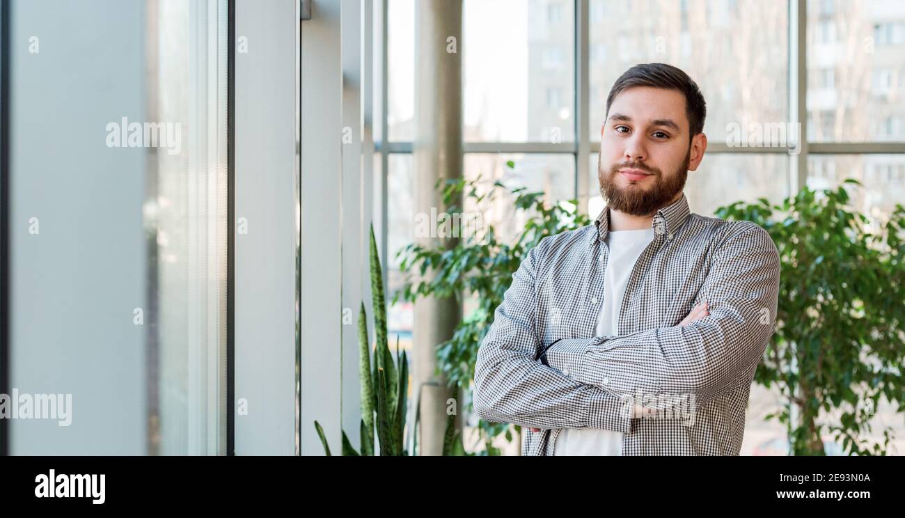Homme caucasien debout près de la fenêtre pleine longueur dans le bureau moderne avec des plantes. Le jeune homme barbu a croisé les bras. Homme d'affaires élégant Banque D'Images