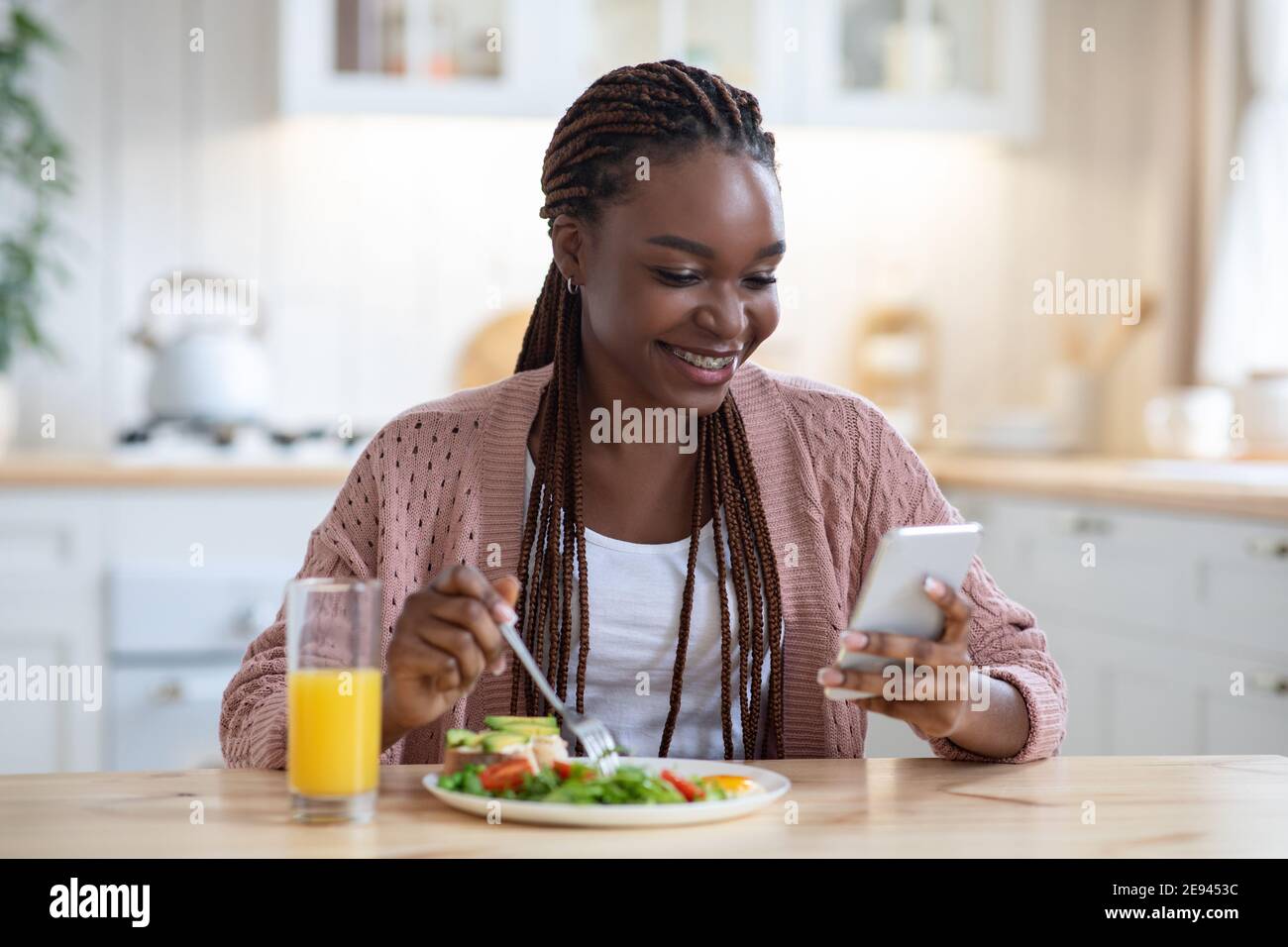 Femme noire souriante utilisant un smartphone tout en prenant le petit déjeuner dans la cuisine à la maison Banque D'Images