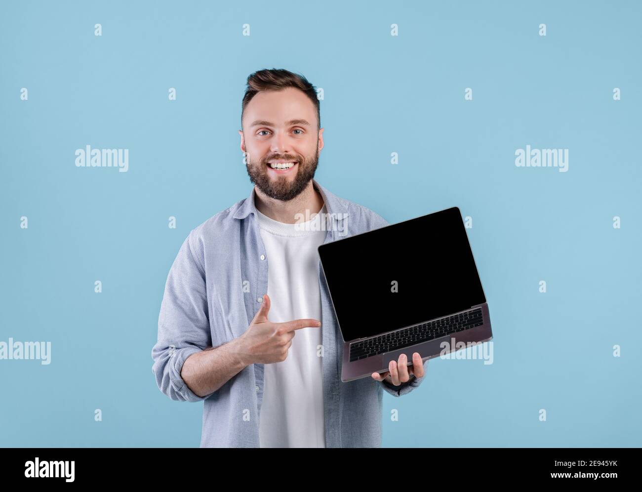 Jeune homme positif pointant à l'écran d'ordinateur portable vide avec de l'espace pour votre conception de site Web sur fond bleu, maquette Banque D'Images